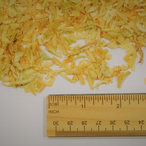 May include: A pile of dried, yellow onion flakes with a wooden ruler for scale. The ruler is marked in inches and shows measurements up to 3 inches. The onion flakes are a light yellow color with some pieces showing a slight orange hue.