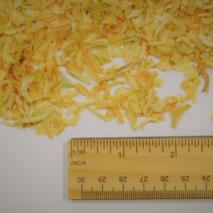 May include: A pile of dried, yellow onion flakes with a wooden ruler for scale. The ruler is marked in inches and shows measurements up to 3 inches. The onion flakes are scattered on a white surface.