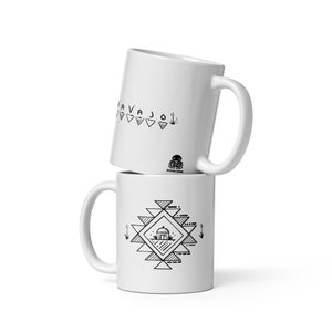 May include: Two white ceramic mugs, one stacked on top of the other. The top mug has the word "NAVAJO" in black, with triangles and other small designs. The bottom mug features a geometric design with a small house in the centre, also in black.