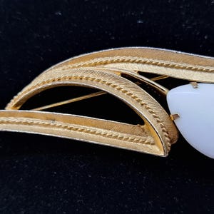 May include: A gold-toned brooch with a leaf design and a white, teardrop-shaped stone. The brooch has a textured finish and a pin closure on the back. The design is reminiscent of a stylized flower or leaf.