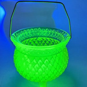 May include: A bright neon green glass cauldron featuring a textured diamond pattern across its rounded body and rim. The vessel is supported by three small glass feet and includes a thin, arched metal handle attached to the top edge.
