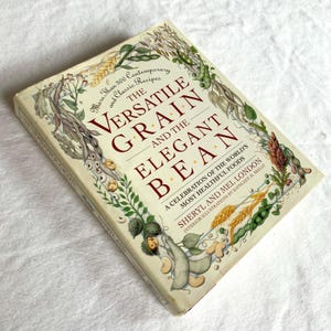 May include: A hardcover cookbook titled "The Versatile Grain and the Elegant Bean" with a cream-colored cover. The title is in large, maroon letters. The cover features illustrations of grains and beans, with text that reads "More Than 300 Contemporary and Classic Recipes."
