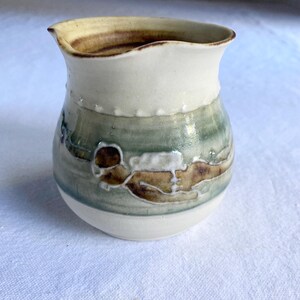 May include: A handmade ceramic pitcher with a cream-colored top and base, and a green and brown patterned body. The pitcher has a unique, organic shape and a slightly flared rim. The design features abstract shapes and textures.