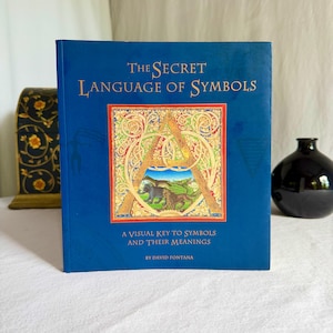 May include: A hardback book with a blue cover, titled "The Secret Language of Symbols". The cover has gold lettering and a decorative "A" with an illustration. The book is by David Fontana and explores the meaning of symbols.