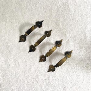 Puede incluir: Cuatro tiradores de cajón antiguos de latón con un asa curvada y terminales decorativos. Los tiradores son de un color latón cálido y envejecido, con un aspecto ligeramente deslustrado. Los terminales tienen forma de lágrima.