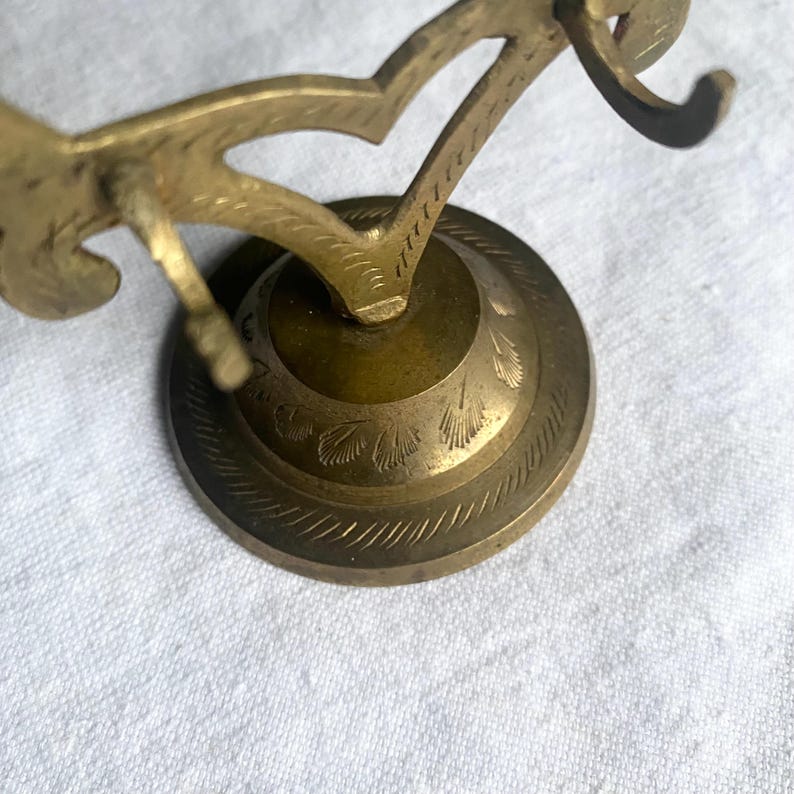 May include: A vintage brass decorative object with an ornate design. The object has a circular base with etched floral patterns and a curved, stylized top. The brass has a warm, aged patina, suggesting its vintage origin.