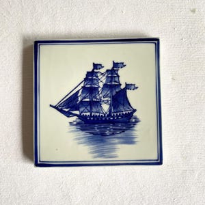 May include: Square ceramic tile featuring a blue and white illustration of a tall ship sailing on water. The ship has multiple masts and sails, with flags at the top. The tile has a blue border and a glossy finish.