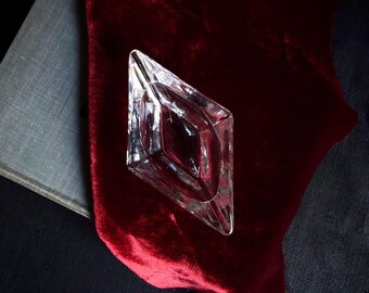 May include: A clear, diamond-shaped glass dish with faceted edges and a recessed center. The object features a transparent, geometric design with sharp, pointed ends and a smooth, reflective surface, showcasing a clean and minimalist aesthetic.