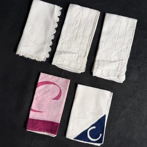 May include: Five folded rectangular napkins. One is white with a lace edge, two are white with embroidered grid patterns, one is pink with a dark pink curved design, and one is white with a dark blue corner triangle featuring a white letter C.