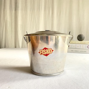 May include: A vintage metal bucket with a lid and handle. The bucket has a silver finish and a red and yellow "FISKO" logo. The bucket is sitting on a white surface.
