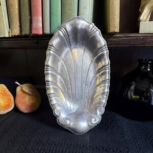May include: An oval silver-toned metal dish featuring a decorative shell-like design with embossed radiating fluted lines and scrolled details at the base. The surface has a polished metallic finish with subtle reflections throughout the structure.