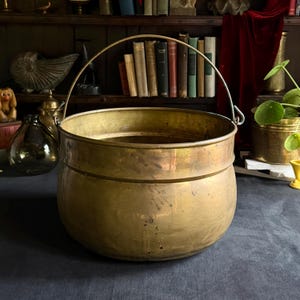 May include: A large, round, vintage-style brass bucket featuring a smooth, polished surface with subtle signs of wear and a patina. The vessel includes a sturdy, arched metal handle attached to two side loops, creating a wide, open-top container.