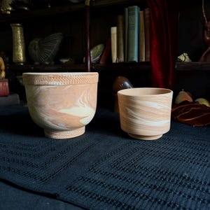 May include: Two ceramic bowls with a marbled pattern of terracotta orange and cream. The larger bowl features a textured, cross-hatched pattern along the rim and base, while the smaller bowl has a smooth, rounded exterior and a raised circular foot.