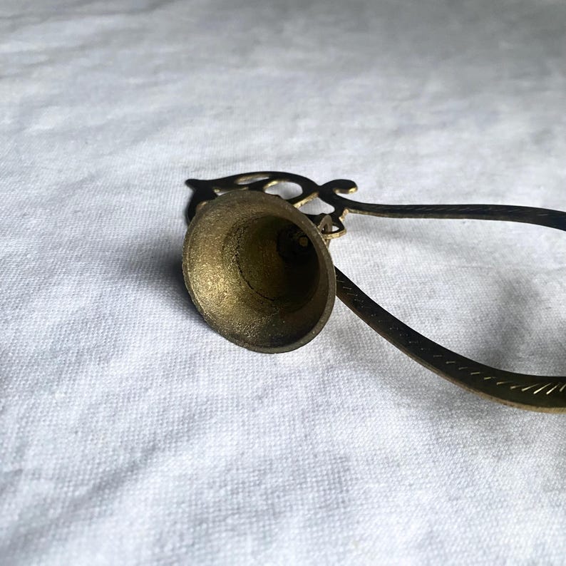 May include: A vintage brass bell with a long handle and ornate detailing. The bell has a flared shape and a small clapper inside. The handle has a decorative design. The bell is likely used for calling or signaling.