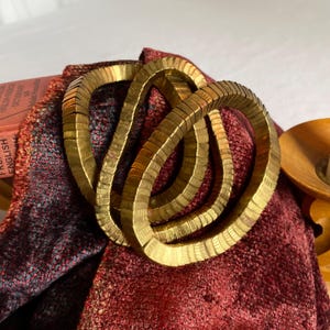 May include: A gold-toned metal bracelet with a knotted design. The bracelet is made of small, rectangular segments linked together. It rests on a textured, red and black fabric, with a book visible in the background. The bracelet's design is a series of interlocking loops.