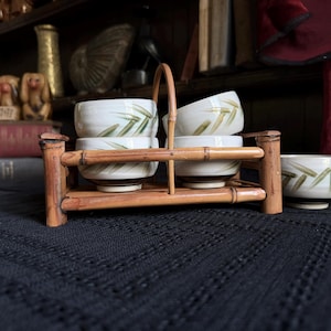 May include: A set of four ceramic bowls with a white base and green bamboo leaf patterns, held in a rectangular bamboo carrier with a central handle. Each bowl features a brown base, and an additional matching bowl sits to the right of the carrier.