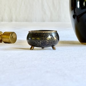 May include: A small, black decorative bowl with a floral pattern and three short legs. The bowl has a gold rim and is likely made of metal. The bowl is a decorative item.