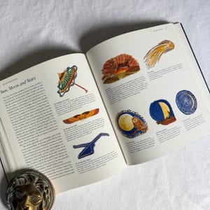 May include: An open book displaying illustrations of celestial bodies. The pages show colourful drawings of a sunshade, a comet, and a full moon with an owl. The book is titled "The Natural World". The book is open on a white surface.