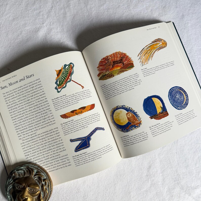 May include: An open book displaying illustrations of celestial bodies. The pages show colourful drawings of a sunshade, a comet, and a full moon with an owl. The book is titled "The Natural World". The book is open on a white surface.