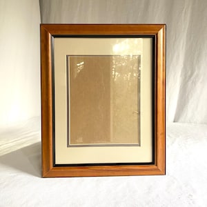 May include: A rectangular wooden picture frame with a warm brown finish. The frame has a black inner border, a cream-colored mat, and a clear glass or acrylic cover. The frame is designed to hold a photograph or artwork.
