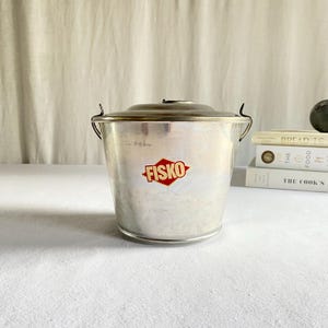 May include: A vintage metal bucket with a lid and handle. The bucket has a red and white "FISKO" logo on the front. The bucket is sitting on a white surface.