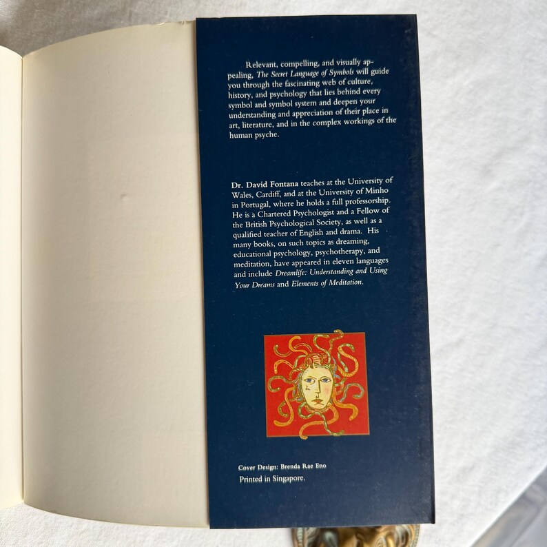 May include: A dark blue book cover with white text, titled "The Secret Language of Symbols". The text describes the book's content and author. A red square with a gold illustration of a face with snakes is featured on the cover.