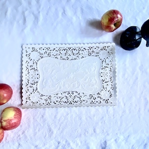 Puede incluir: Un mantel individual blanco rectangular con borde festoneado y calados intrincados. El centro presenta un diseño floral en relieve. Manzanas rojas y amarillas están dispersas alrededor del mantel.