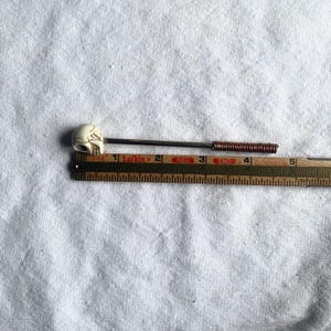 May include: A decorative pin with a skull-shaped head. The pin has a dark metal shaft with a copper coil near the end. The skull is off-white with carved details. The pin is approximately 4 inches long.
