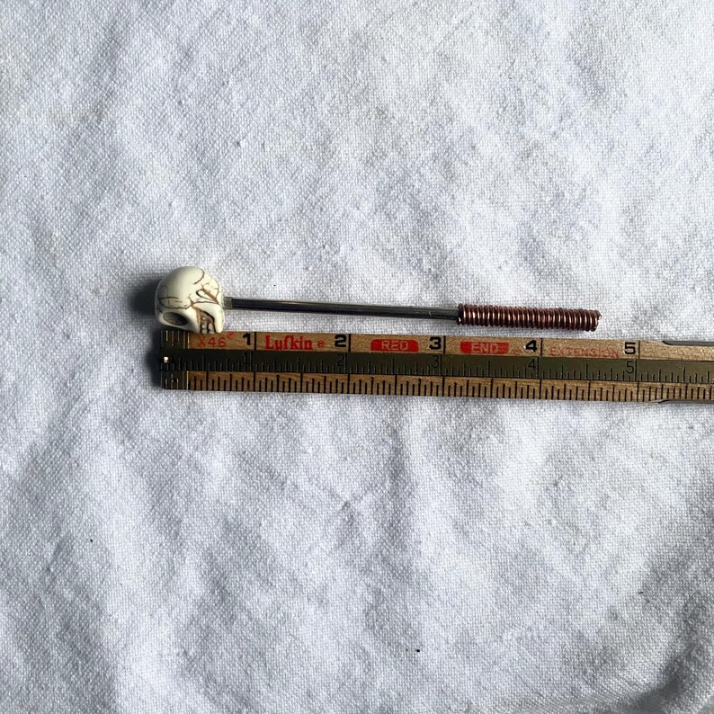 May include: A decorative pin with a skull-shaped head. The pin has a dark metal shaft with a copper coil near the end. The skull is off-white with carved details. The pin is approximately 4 inches long.
