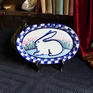 May include: An oval ceramic platter featuring a hand-painted white rabbit in the center, surrounded by green grass and pink flowers. The rim is decorated with a blue and white checkered pattern, and the piece rests on a black display stand.