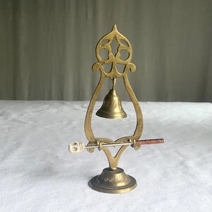 May include: A vintage brass bell with a decorative frame and a small clapper. The bell is mounted on a round base with etched details. A small bone-like object and a copper-colored rod are attached to the frame.