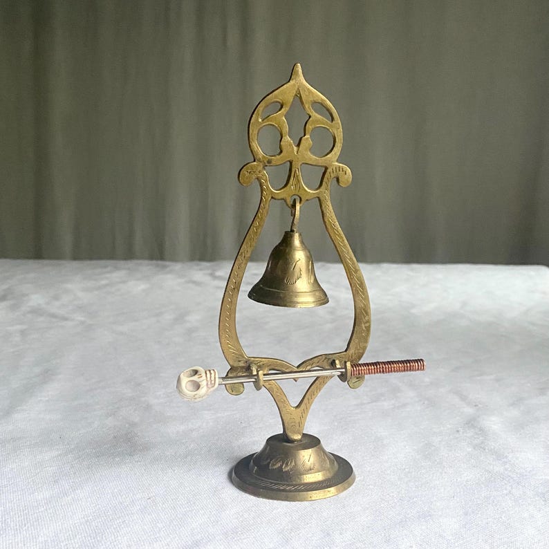 May include: A vintage brass bell with a decorative frame and a small clapper. The bell is mounted on a round base with etched details. A small bone-like object and a copper-colored rod are attached to the frame.