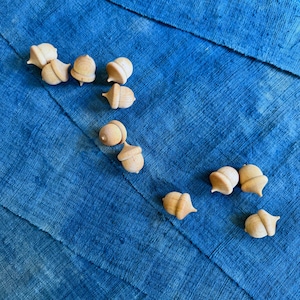 May include: A collection of small, wooden acorn-shaped ornaments scattered on a textured, deep blue fabric. The acorns are light brown with detailed caps and smooth bodies. The fabric has visible seams and a slightly faded appearance.
