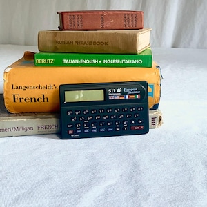 Puede incluir: Una pila de libros antiguos con títulos en inglés, francés, italiano y ruso. Un traductor europeo SII negro está encima de los libros. Los libros son de varios colores, incluyendo rojo, verde y amarillo.