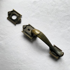 Może przedstawiać: Antyczny mosiężny uchwyt do drzwi i rozeta. Uchwyt ma zakrzywiony kształt z dekoracyjnymi detalami na prostokątnej płycie tylnej. Rozeta to pasująca okrągła płyta z centralnym otworem i ozdobnymi krawędziami. Okucia są prawdopodobnie do drzwi w stylu vintage.