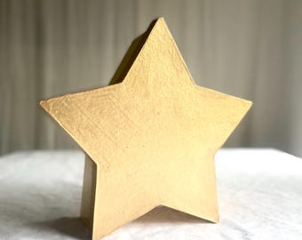 May include: A gold-colored, five-pointed star-shaped decorative object. The star has a textured surface and a three-dimensional design, standing upright on a flat base. The star is likely made of wood or a similar material.