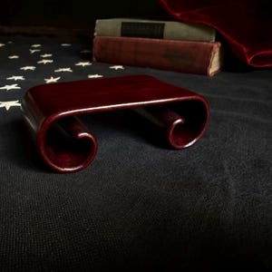 May include: A dark reddish-brown wooden display stand featuring a smooth, rectangular top surface and two curved, scroll-like legs on either side. The stand has a polished finish and is positioned on a dark fabric surface with white star patterns.