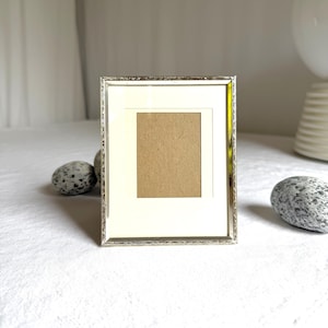 May include: A silver picture frame with a decorative edge, cream mount, and brown backing. The rectangular frame is designed for a photograph. Two speckled stones are placed near the frame. The frame is approximately 15 cm in height.