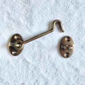 May include: A brass-colored hook and eye latch set. The set includes an oval base with three screw holes, a straight arm, and a curved hook. The matching eye piece has an oval base with two screw holes. The hardware is designed for securing doors or windows.