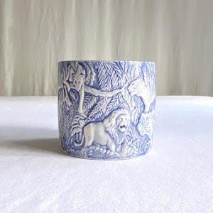 Puede incluir: Jardinera de cerámica azul y blanca con una escena de la jungla. La jardinera presenta imágenes en relieve de leones, monos y follaje. El diseño es detallado y la jardinera tiene forma cilíndrica.