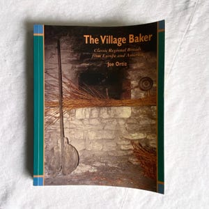 Może przedstawiać: Książka zatytułowana "The Village Baker" z okładką przedstawiającą wnętrze rustykalnego kamiennego pieca. Tekst na okładce zawiera "Classic Regional Breads from Europe and America" oraz nazwisko autora, Joe Ortiz. Książka ma turkusową i pomarańczową obwódkę.