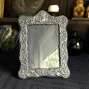 May include: A rectangular picture frame with a metallic silver finish and an ornate, raised relief border featuring intricate floral and scrollwork patterns. The frame has a central glass pane and a scalloped top edge, showcasing a detailed design.