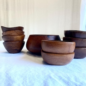 Könnte beinhalten: Eine Sammlung von Holzschalen in verschiedenen Größen und Formen. Die Schalen sind aus dunkelbraunem Holz gefertigt und haben eine glatte, polierte Oberfläche. Einige Schalen sind gestapelt, andere werden einzeln präsentiert. Die Schalen sind auf einer weißen Oberfläche angeordnet.