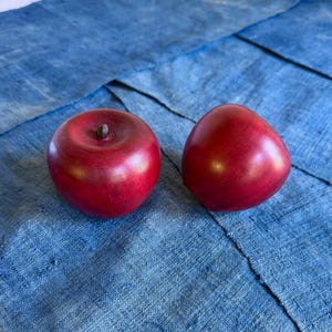 May include: Two red wooden apples with a glossy finish rest on a blue fabric surface. One apple has a small, dark stem. The apples are a vibrant red color, and the fabric has a textured appearance.