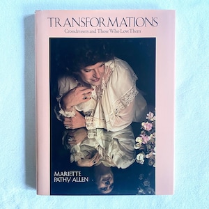 Puede incluir: Un libro titulado "Transformations: Crossdressers and Those Who Love Them" con una portada rosa. La portada presenta una fotografía de una persona con una prenda blanca con encaje, reflejada en una superficie oscura. El nombre del autor es visible.