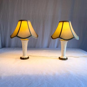 May include: A pair of vintage table lamps. Each lamp features a white, floral-patterned base, a yellow lampshade with a blue-green trim, and a bronze-coloured base. The lamps are illuminated, emitting a warm light.
