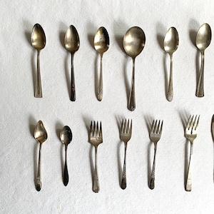 Puede incluir: Una variedad de cubiertos antiguos, que incluyen cucharas y tenedores, dispuestos sobre una superficie blanca. Las cucharas varían en tamaño y forma, algunas con detalles ornamentales en los mangos. Los tenedores tienen púas y elementos decorativos. Los cubiertos tienen un aspecto antiguo y deslustrado.