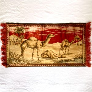 May include: A rectangular tapestry featuring a desert scene with camels, palm trees, and buildings. The background is a deep red, and the tapestry has a brown border and red fringe.