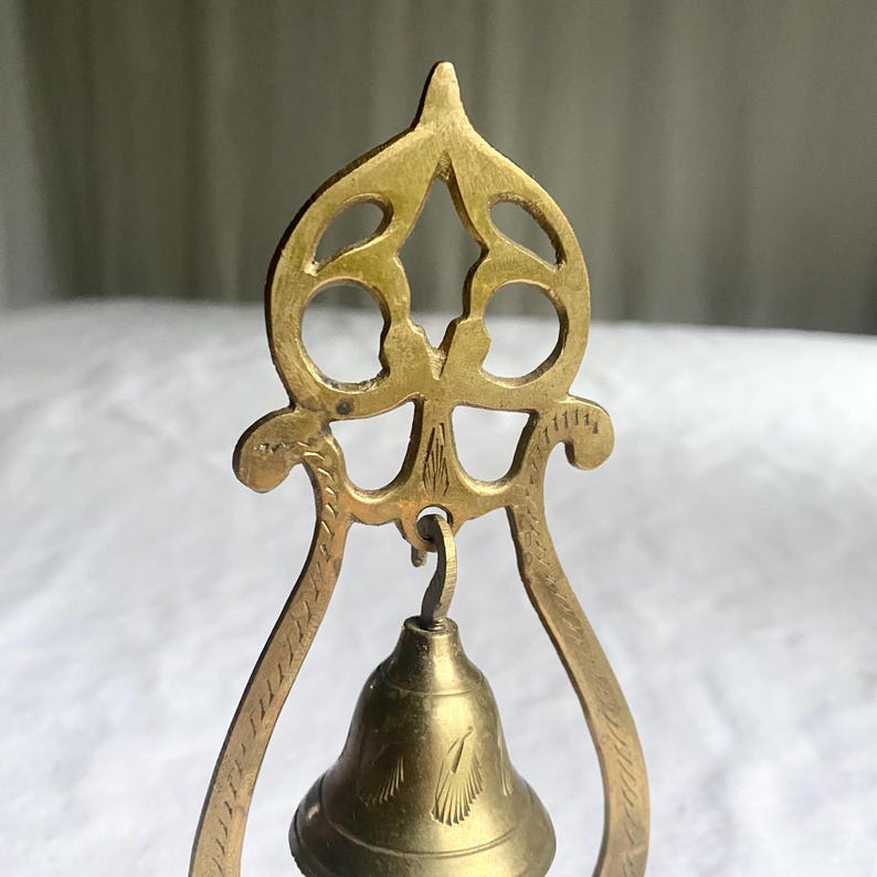 May include: A close-up of a brass bell with an ornate, openwork top and a small bell hanging below. The bell has etched details and a warm, golden hue. The frame has curved sides with etched patterns.