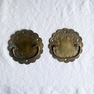 May include: Two antique brass drawer pulls. Each pull has a scalloped edge and a floral design. The pulls have a curved handle and are mounted with small screws. The brass has a slightly aged patina.